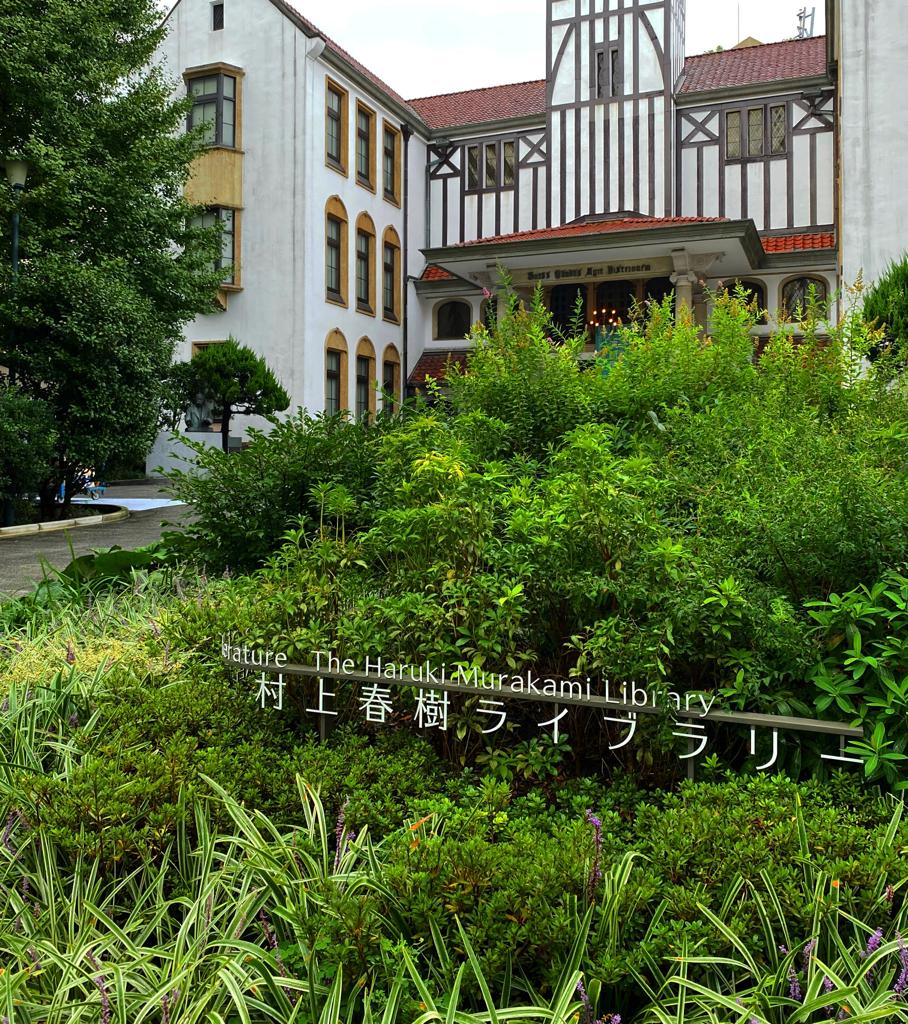 Photo of Haruki Murakami Library signage in the foreground surrounded by grass and bush; and Tsubouchi Memorial Theatre in the background. The facade of the theatre is in Shakespearean style.